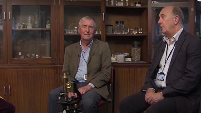  Professors Geoff Gill and Nick Beeching being interviewed in the laboratory in front of a display cabinet of historical laboratory equipment and samples 